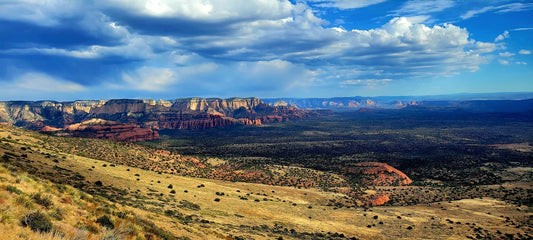 Wide desert landscape of the Verde Valley under midday light—red rock cliffs, sparse green vegetation, and a bright blue sky with drifting clouds.
